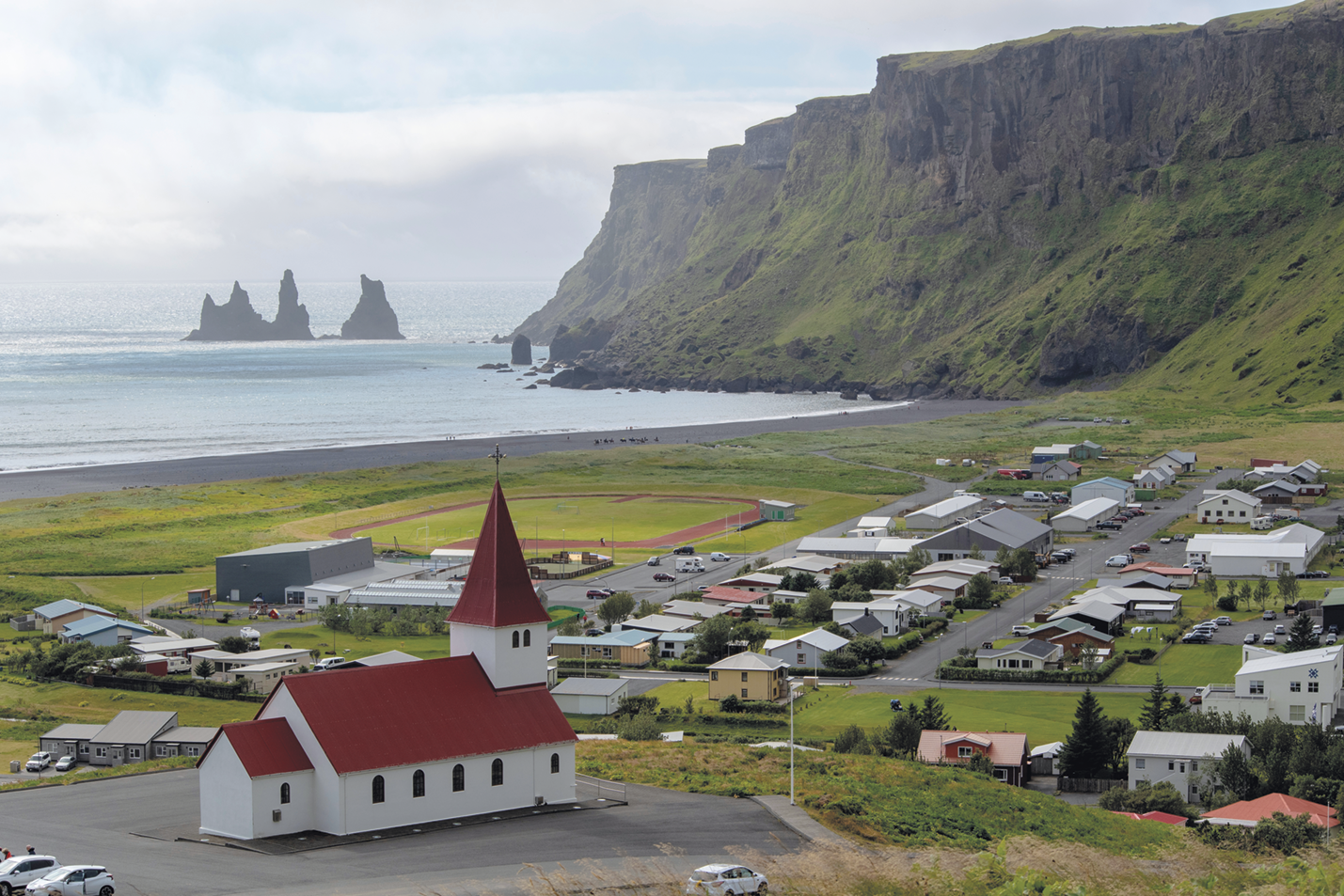 Fotografia. Vista do alto. À esquerda, uma pequena igreja com telhado vermelho, em frene a ela, casas divididas em quarteirões. À esquerda, o mar com formações rochosas e à direita, montanhas cobertas por vegetação.