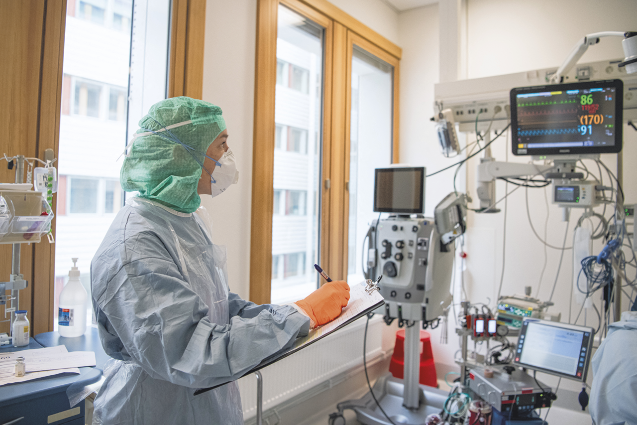 Fotografia. Uma pessoa usando touca, máscara, avental e luvas, olhando um monitor com ondas e escrevendo em uma prancheta. Há diversos equipamentos em frente.