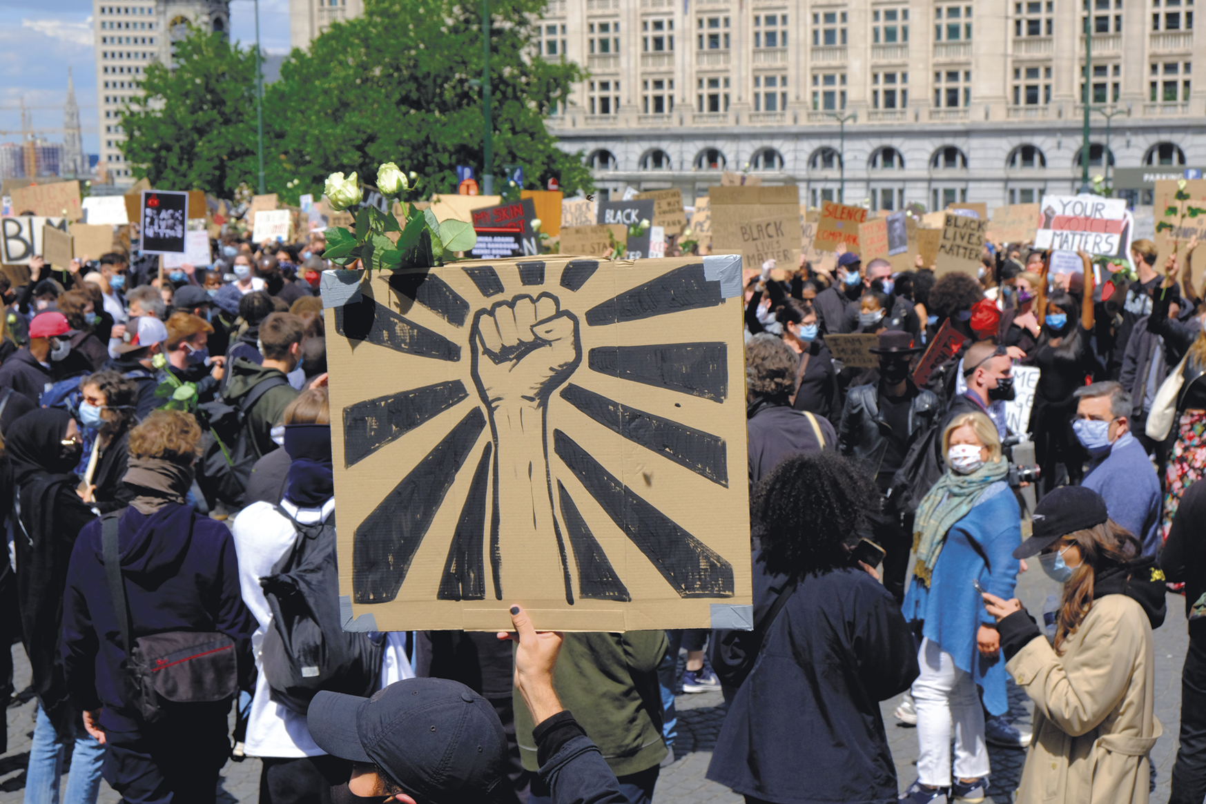 Fotografia. Manifestação com diversas pessoas, com destaque para um cartaz com um punho fechado. Ao fundo, árvores e construções.