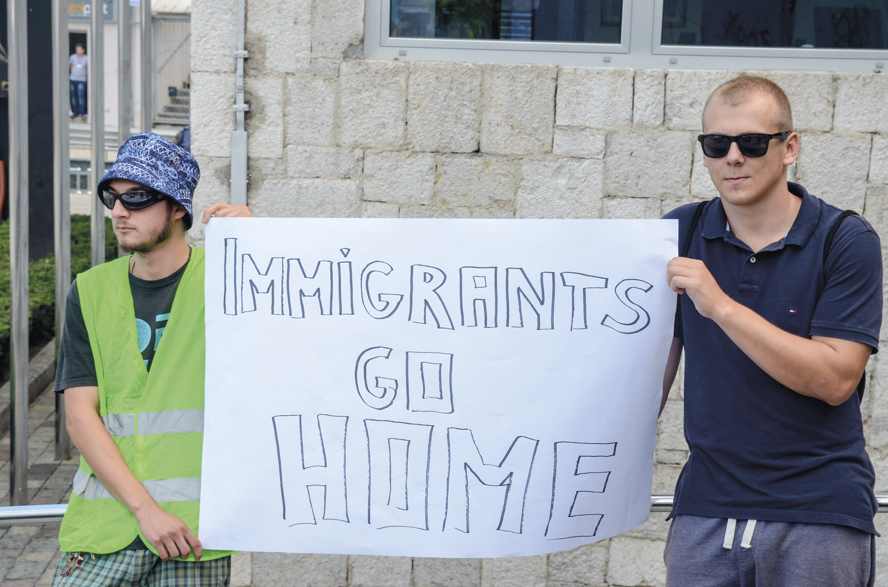 Fotografia. Dois homens brancos em pé, segurando um cartaz com a frase: IMMIGRANTS GO HOME.