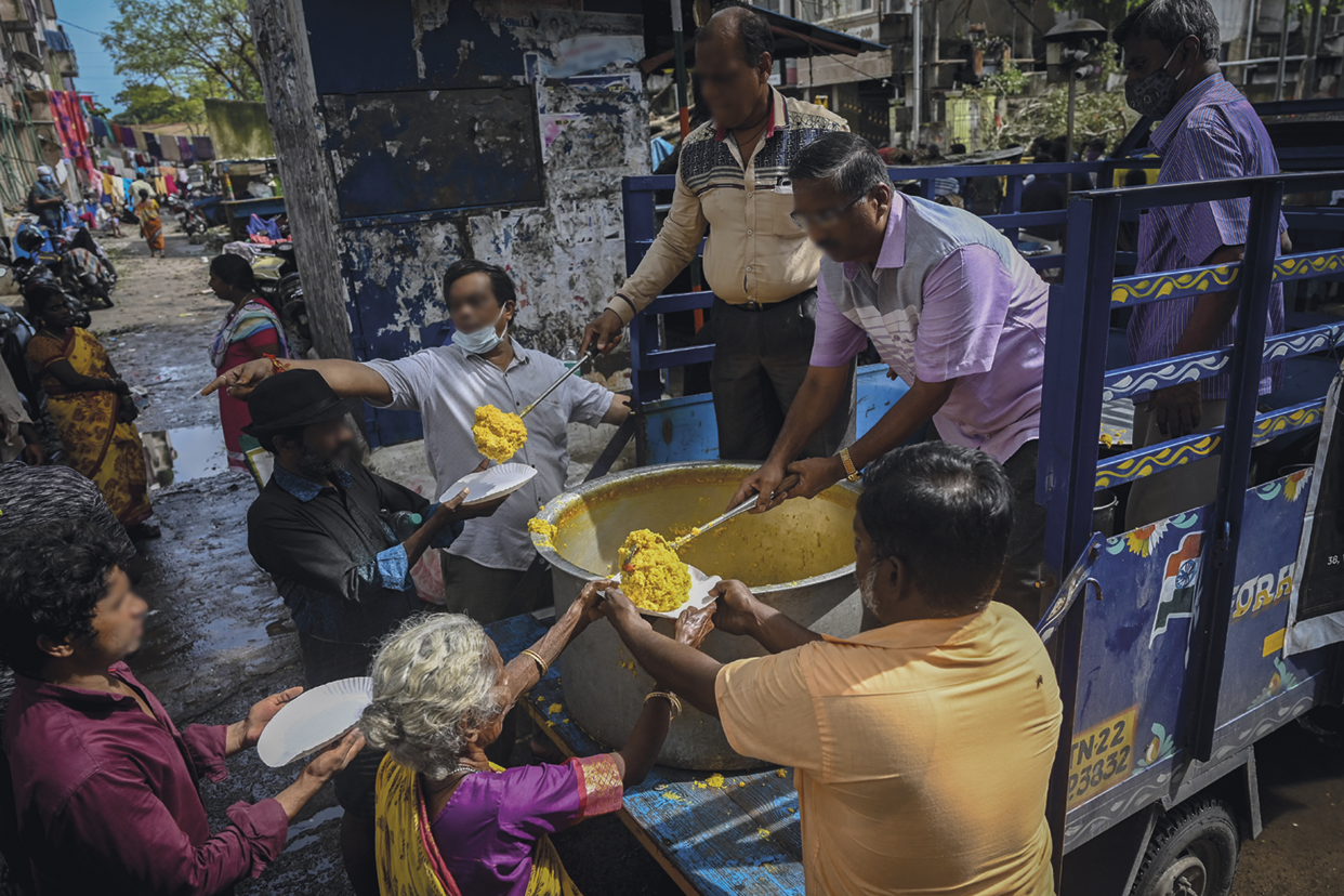 Fotografia. Algumas pessoas formando uma fila extensa em frente a pessoas com panelas que lhes entregam comida. As pessoas na fila estendem pratos.