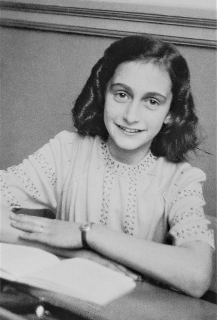 Fotografia em preto e branco. Anne Frank, menina de cabelos na altura dos ombros, sorrindo com os braços sobre uma mesa, em frente a um livro.