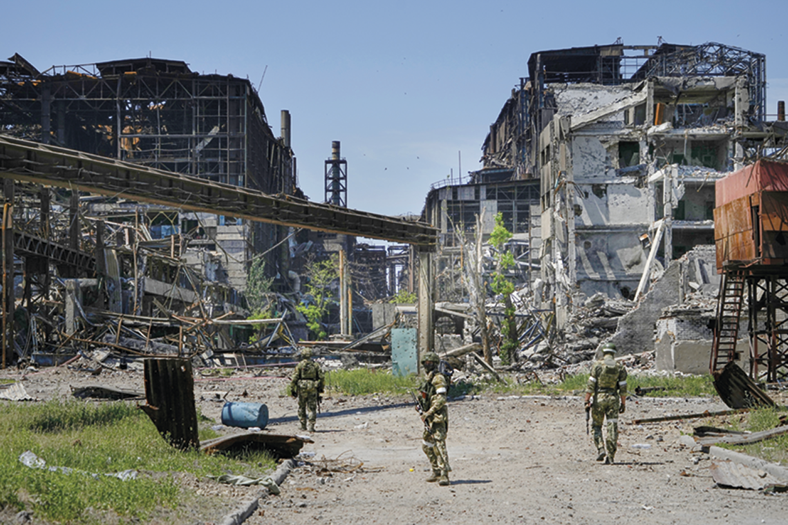 Fotografia. Soldados fardados e armados em meio a uma cidade em ruínas com prédios e estruturas destruídas.