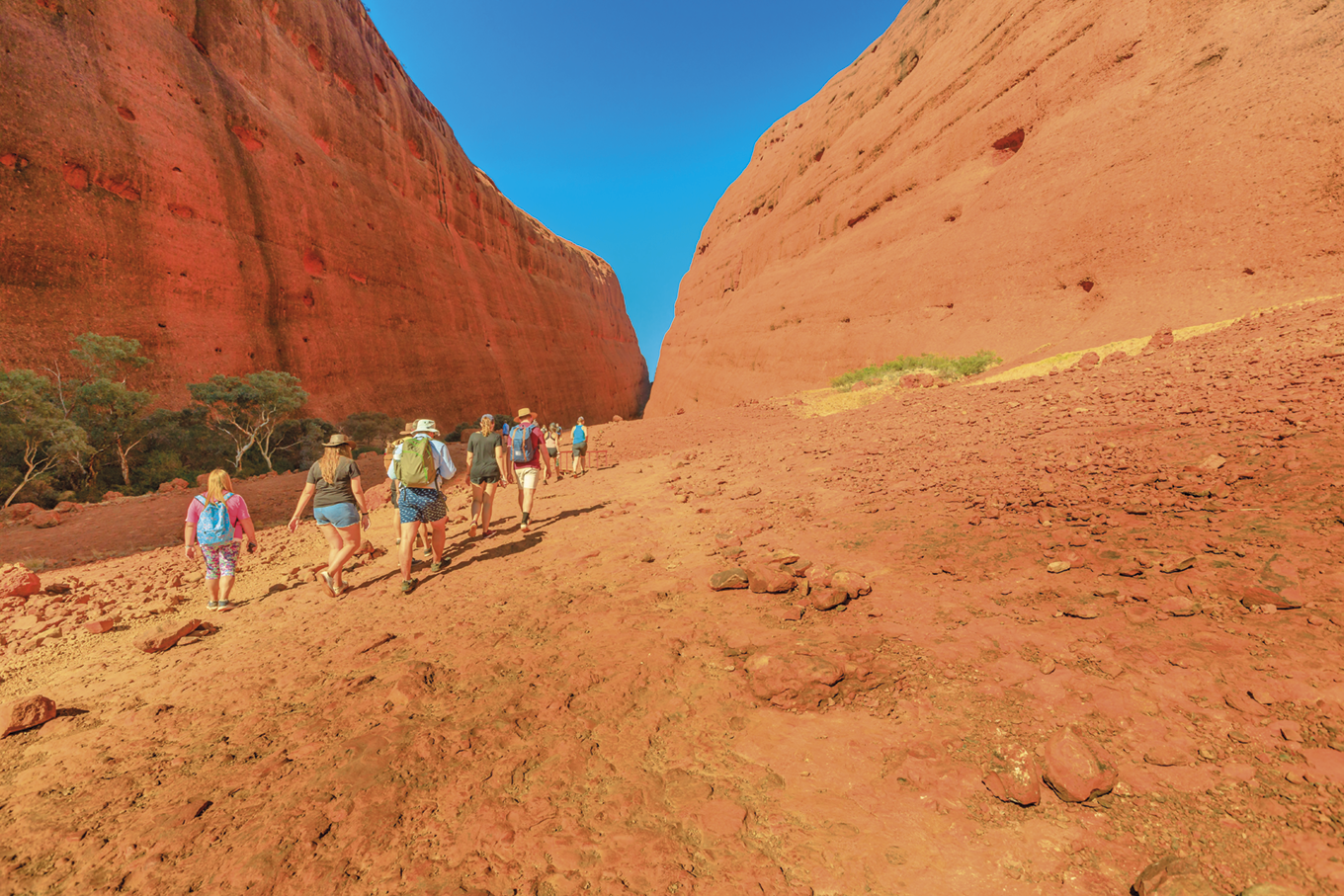 Fotografia. Pessoas caminhando em área deserta com grandes formações rochosas sedimentares.