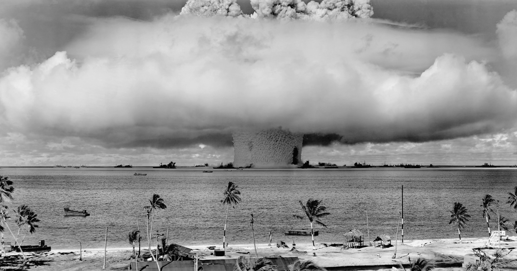 Fotografia em preto e branco. Uma grande explosão no mar. Há uma imensa nuvem de fumaça acima, em formato de cogumelo, em frente há uma praia com coqueiros.