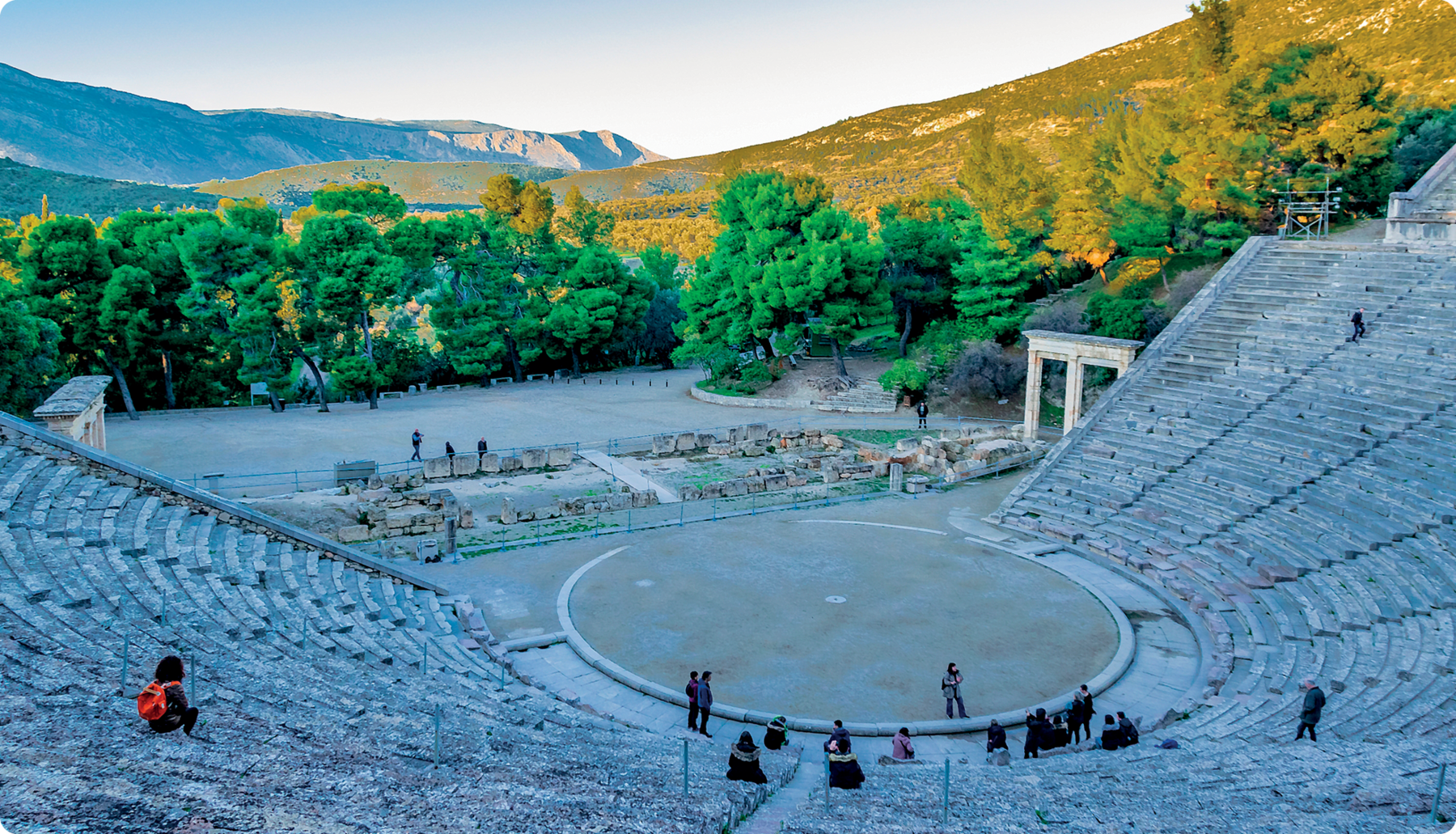 Fotografia. Ruína de um teatro grego. Construção de pedra, sem cobertura e em formato semicircular com vários andares de assento. No centro, um círculo.