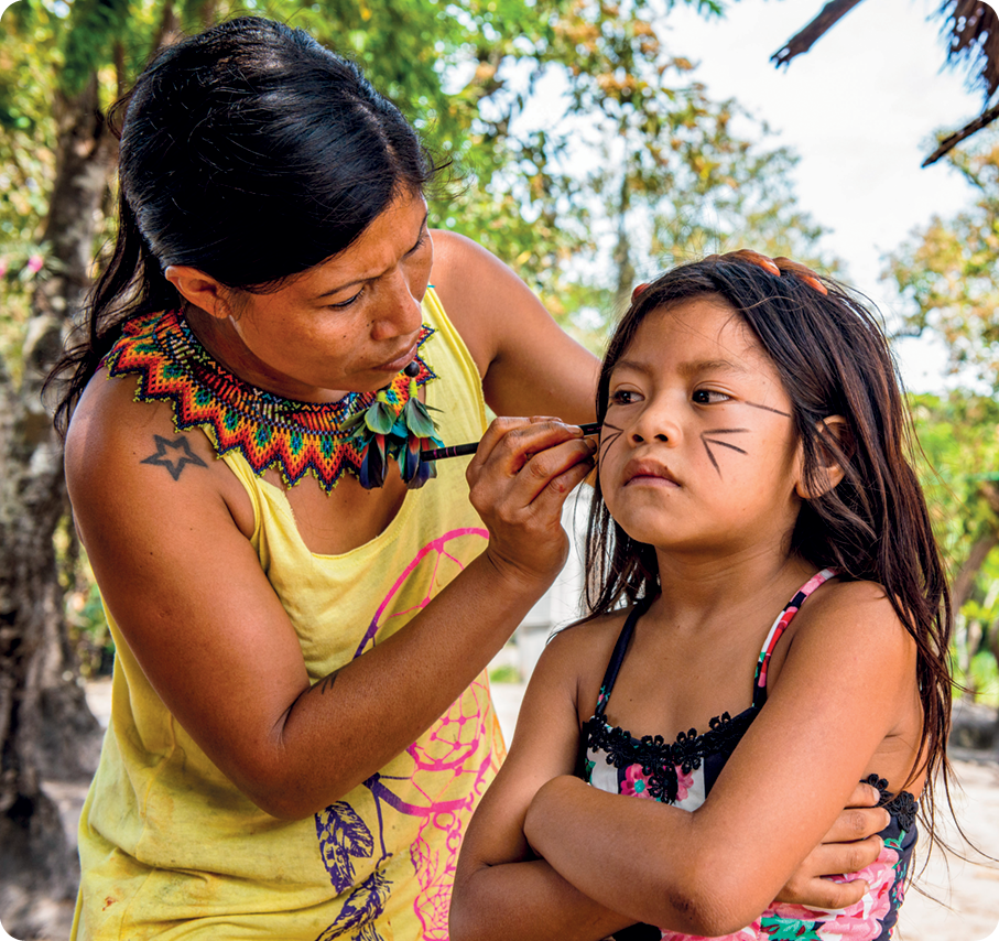 Fotografia. Uma mulher indígena com cabelos presos, vestindo uma roupa amarela e adereços indígenas. Ela está  segurando com uma das mãos um objeto semelhante a um lápis ou uma caneta de cor preta. Ao lado, uma menina indígena com a face sendo pintada pela mulher.