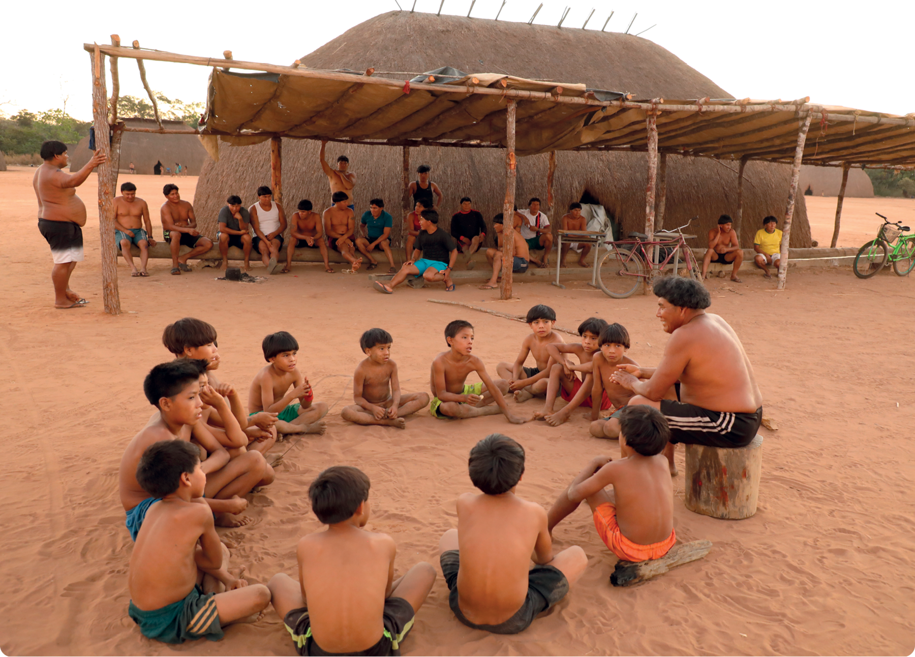 Fotografia. Um homem indígena sentado em um tronco explicando algo. Ao seu redor, um grupo de crianças o escuta atentamente. No fundo, adultos indígenas sentados. Atrás, uma moradia indígena.