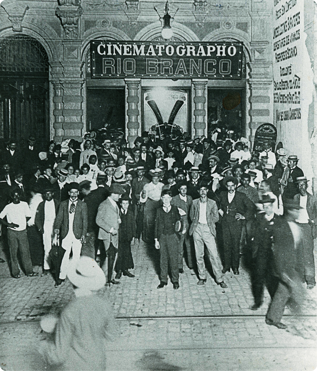Fotografia em preto e branco. Várias pessoas em pé, muitas delas vestindo terno e chapéu. Atrás, cartaz com o texto: CINEMATOGRAPHO.