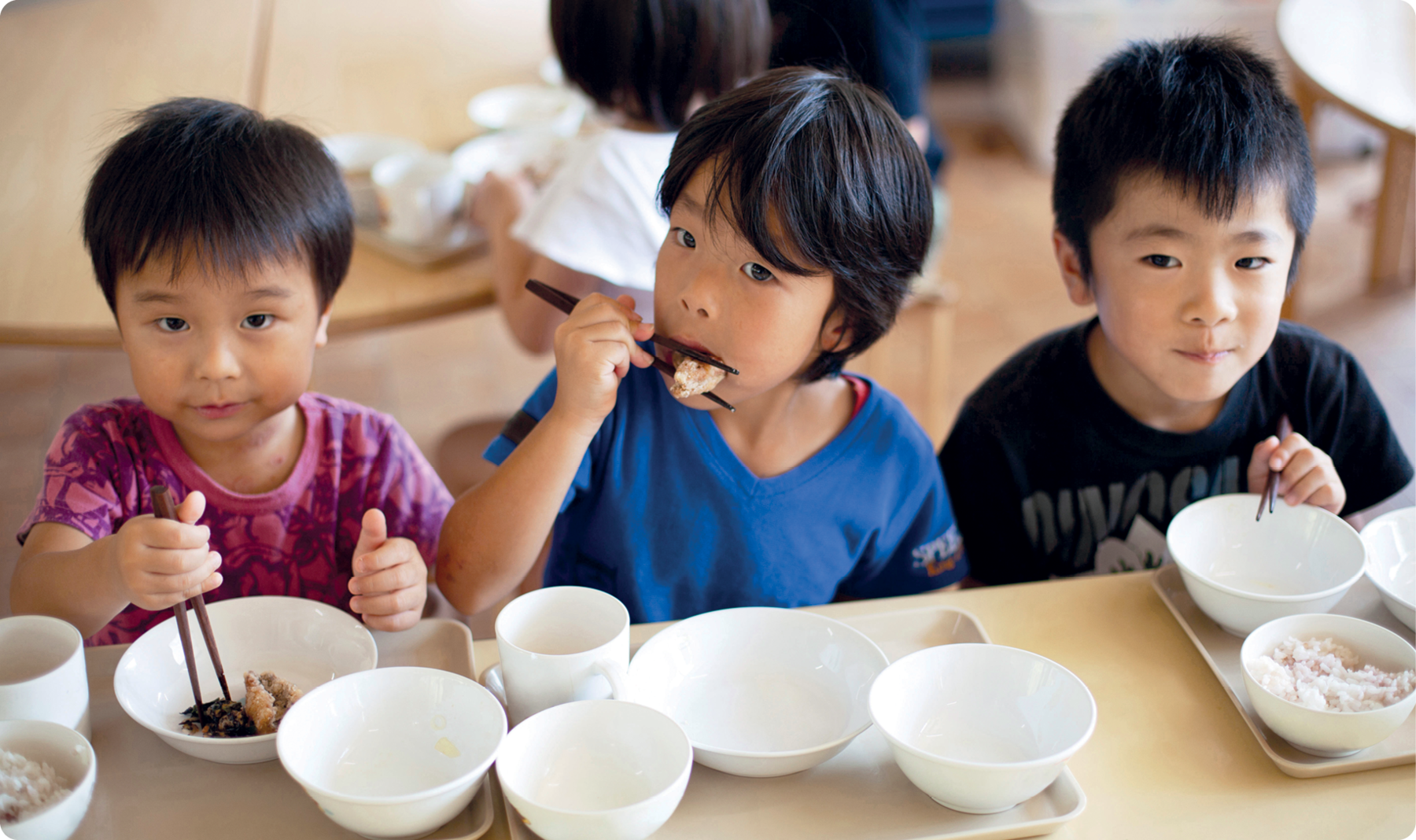 Fotografia. Três crianças japonesas, uma menina com cabelos lisos, usando blusa estampada. Ao lado, um menino usando blusa azul e outro menino usando blusa preta. Elas estão sentados, segurando com as mãos um hashi, duas varetas utilizadas como talheres. Na mesa, potes brancos com alimentos.