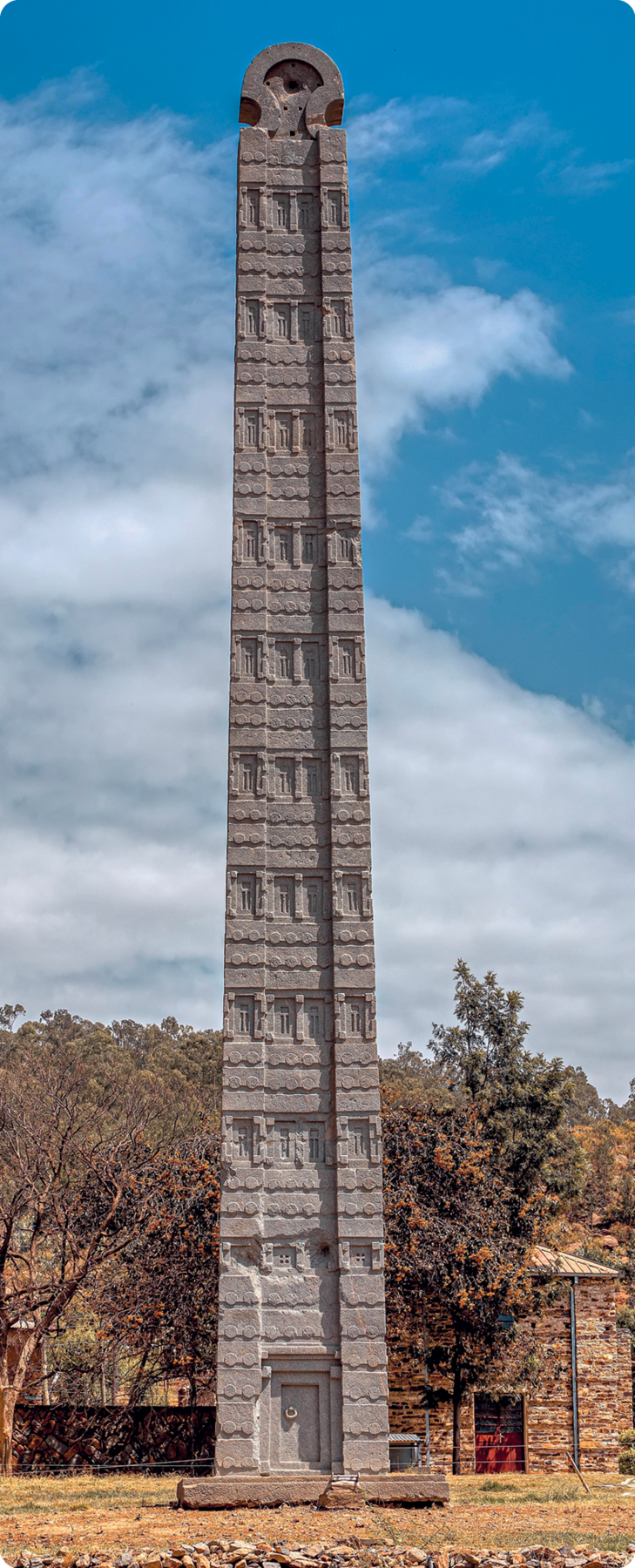Obelisco. Escultura de pedra vertical com base quadrangular, contendo alguns detalhes em relevo. Atrás, algumas árvores.