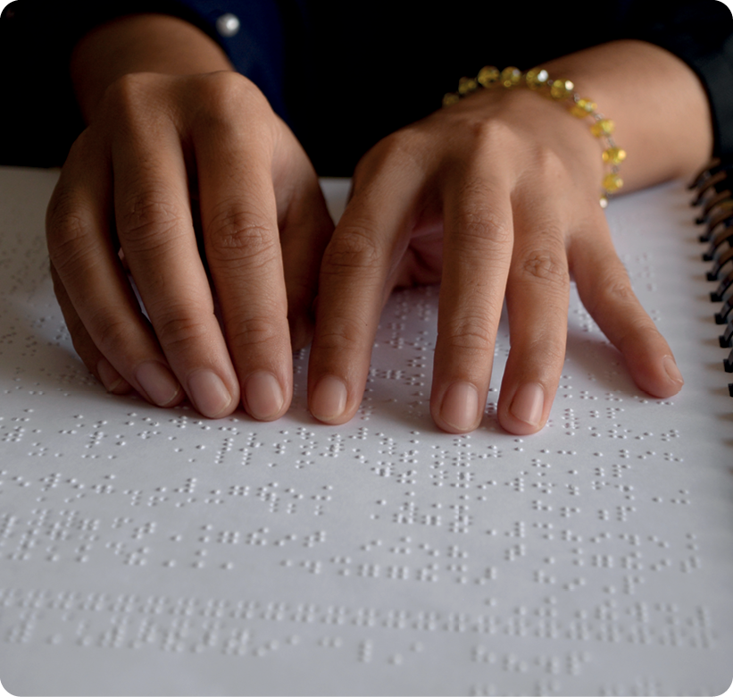 Fotografia. Destacando as mãos de uma pessoa sobre uma folha com texto escrito em braile.