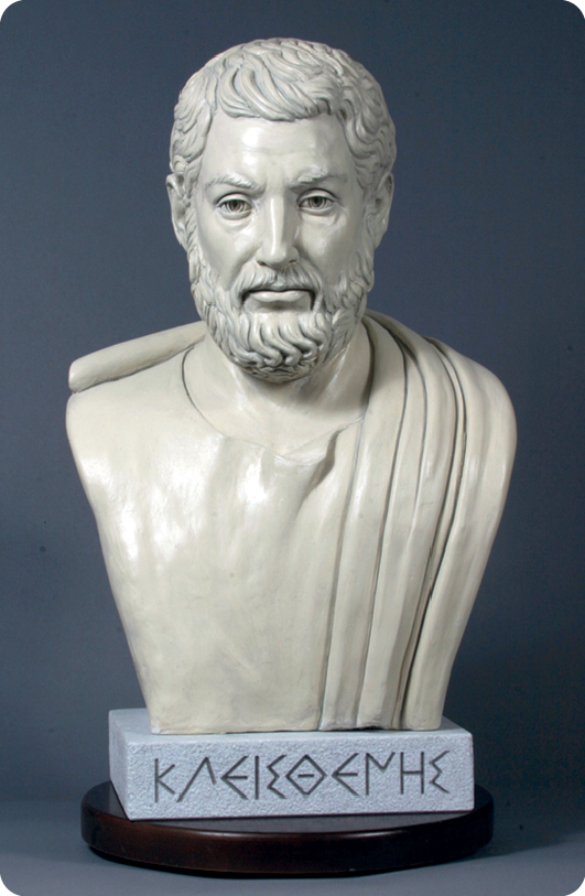 Escultura. Destacando o busto de um homem com cabelos curtos e barba. Abaixo do busto há uma placa com elementos da escrita grega.