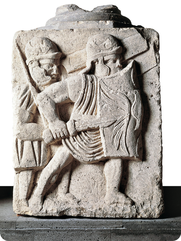 Relevo. Duas figuras humanas usando capacete. A figura da direita segura com as mãos uma espada e um escudo.