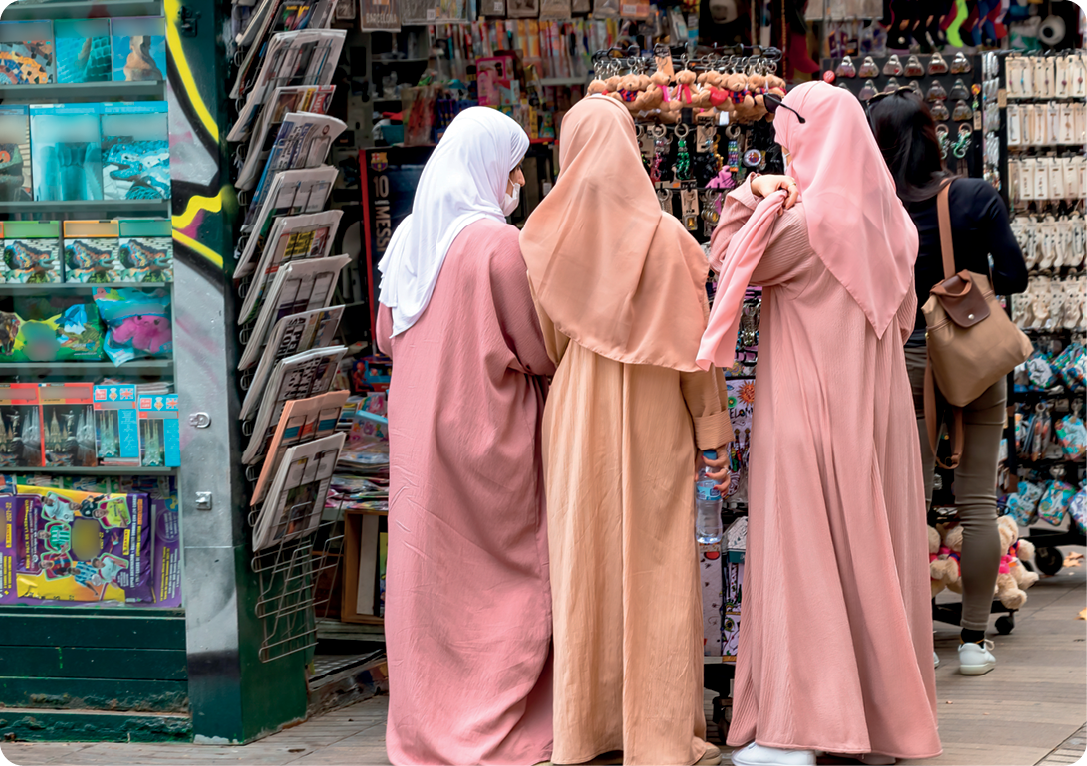 Fotografia. Três mulheres com cabeças e corpos cobertos. Elas estão de pé e de costas, olhando objetos pendurados em uma banca de revistas e jornais.