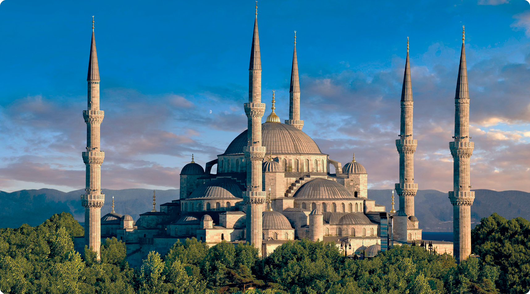 Fotografia. Mesquita composta por uma cúpula grande central, ao redor, cúpulas menores. Nas laterais, cinco grandes colunas ornamentadas. Ao redor, copas de árvores.