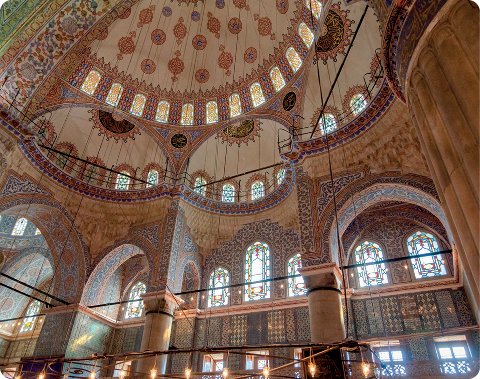 Fotografia. Vista interna do teto alto de uma mesquita. Decoração formada por linhas tracejadas e pintadas nas cores amarelo, azul e vermelho.