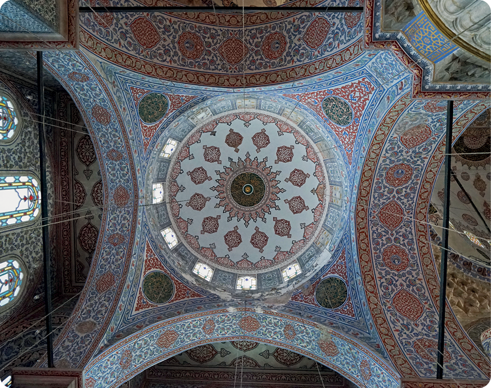Fotografia. Vista interna do teto alto de uma mesquita. Decoração formada por linhas tracejadas e pintadas com as cores azul e vermelho.