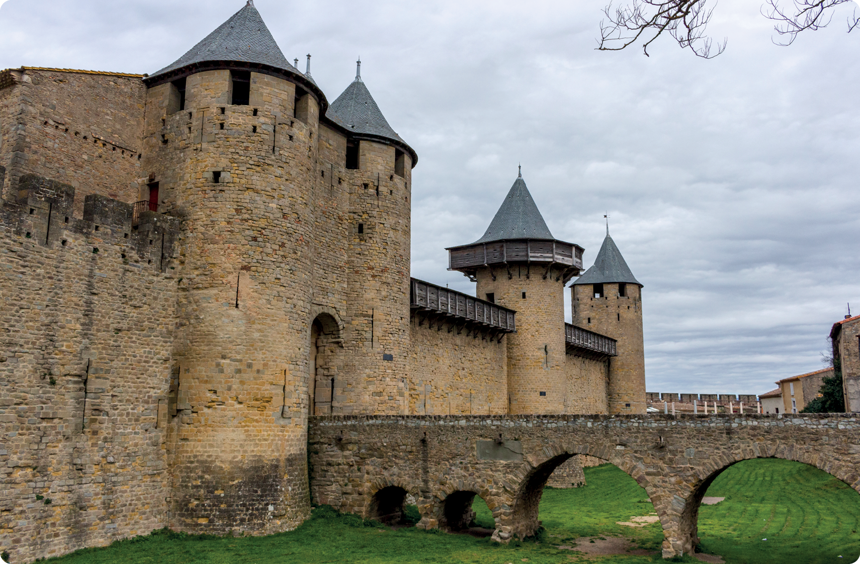 Fotografia. Fachada de castelo de pedra com quatro torres. Em cada torre há uma estrutura no topo em formato de um cone. Na entrada principal, há uma ponte de pedra.