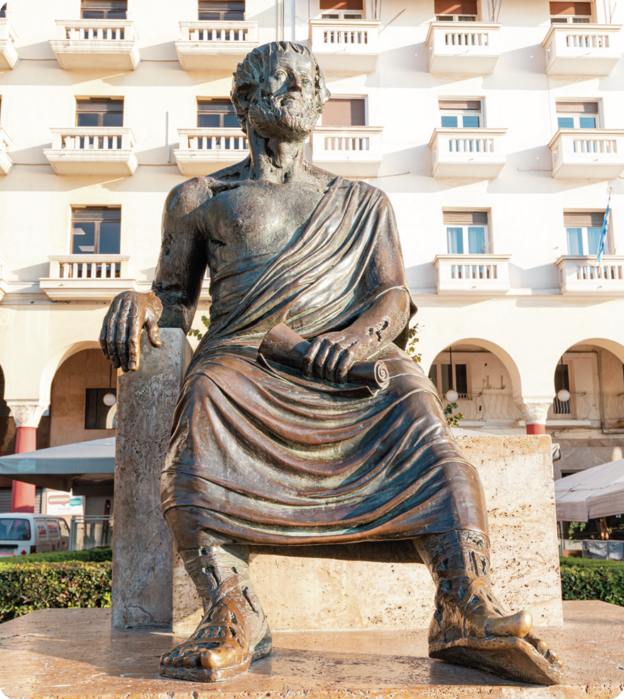 Escultura. Aristóteles, homem com cabelos curtos e barba, tecido ao redor do corpo. Ele está sentado, segurando um objeto com as mãos.