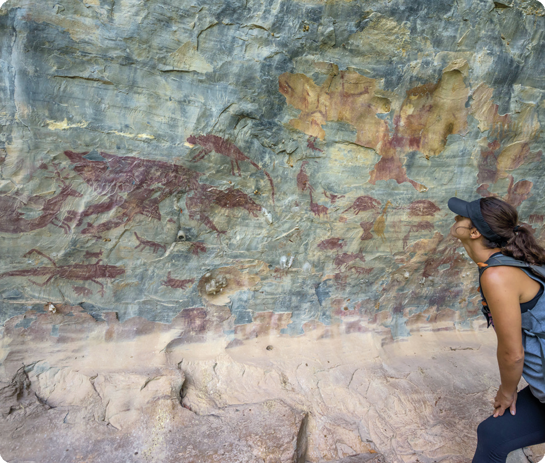 Fotografia. Parede rochosa com pinturas rupestres de silhuetas de animais em cor vermelha. Ao lado, 
uma mulher usando chapéu, está inclinada, com as mãos apoiadas na perna, olhando para as pinturas na rocha.