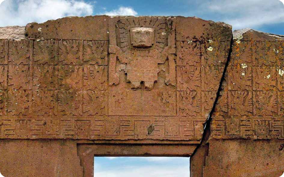Monumento. Parte superior de um portal de pedra com detalhes em relevo de uma pessoa com rosto arredondado, corpo arredondado e raios ao redor de sua cabeça. Nas laterais, detalhes em relevo de quadrados com diversos desenhos.