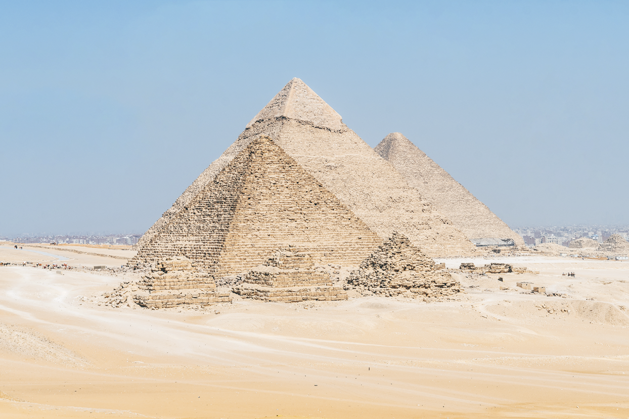 Fotografia. Imagem de seis pirâmides localizadas em uma área deserta, sendo três grandes e três pequenas.