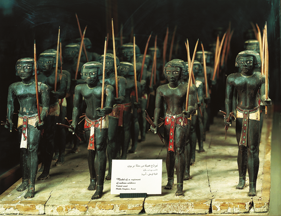 Fotografia. Esculturas de madeira de coloração escura representando figuras masculinas com tecidos coloridos ao redor da cintura e segurando um arco com uma das mãos.