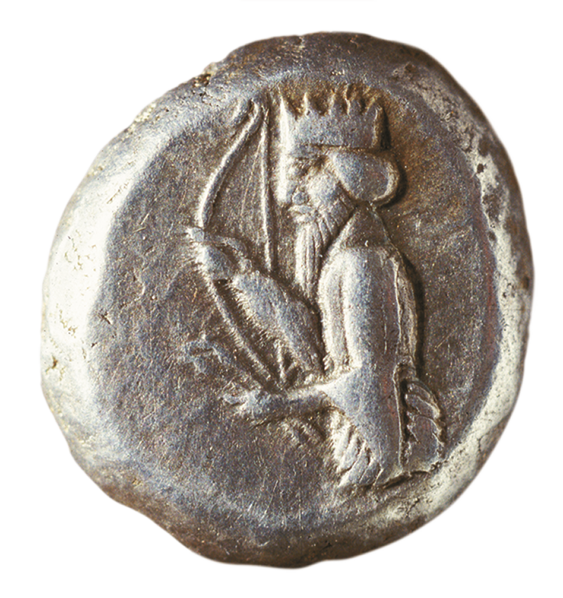 Fotografia. Verso de moeda de prata com detalhes em relevo de um homem usando coroa e segurando com as mãos um arco e flechas.