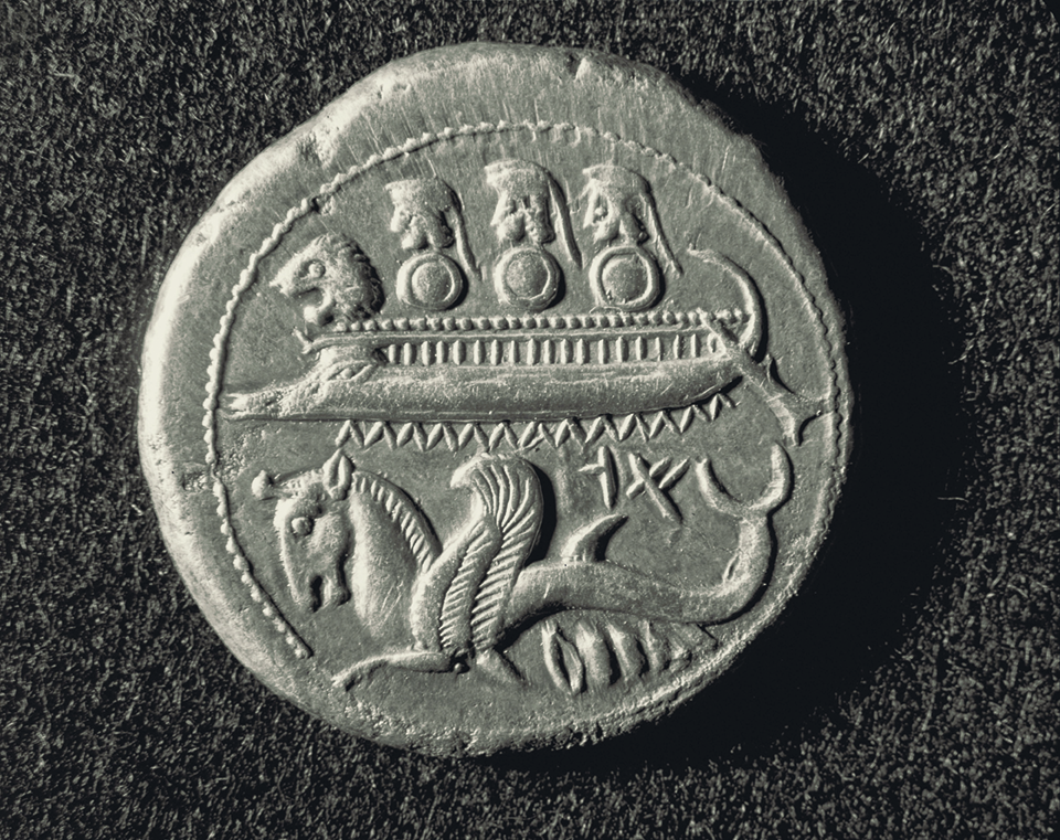 Fotografia. Verso de moeda, na parte superior, representação de três capacetes e escudos de soldados dentro de um navio. Abaixo, um animal mitológico, composto por cabeça de cavalo, asas e cauda de peixe.