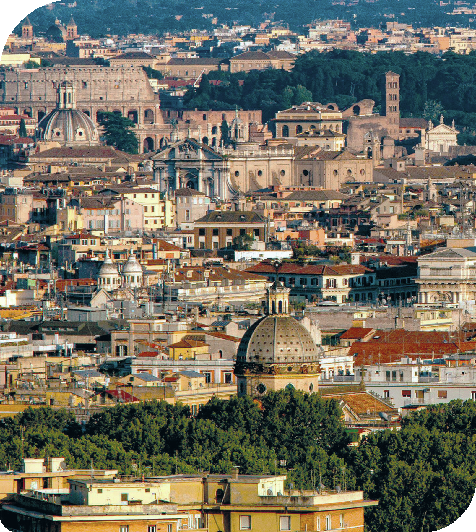 Fotografia. Vista de uma cidade com várias construções grandes e pequenas. No primeiro plano, teto de um prédio com algumas janelas de vidro. Ao centro, algumas construções grandes com cúpulas no telhado e construções menores. Atrás, parte do Coliseu de Roma.