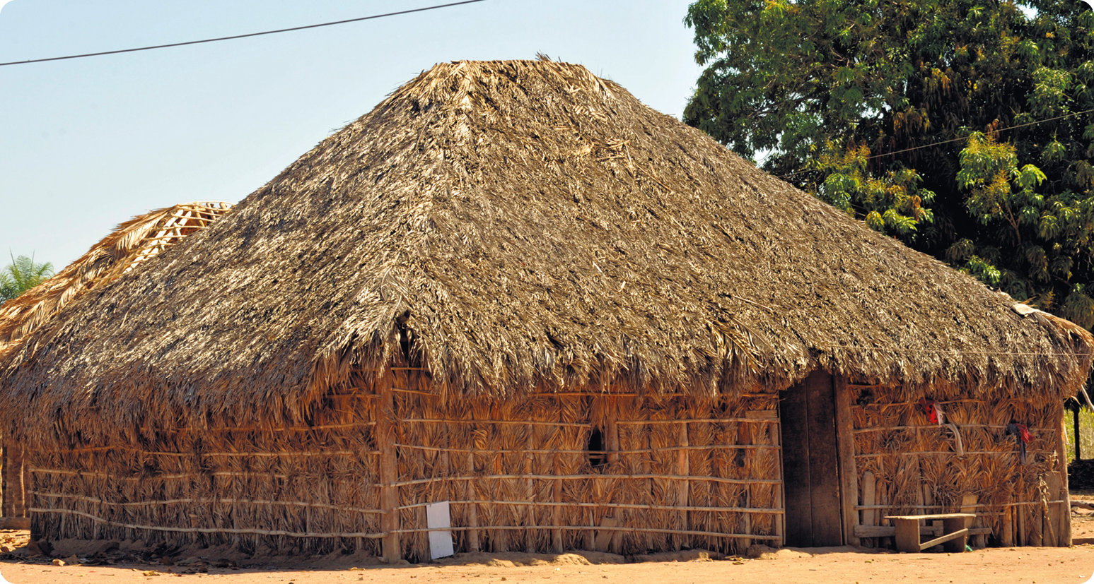 Fotografia. Uma moradia indígena com base retangular e teto em formato triangular feita de madeira e palha.
