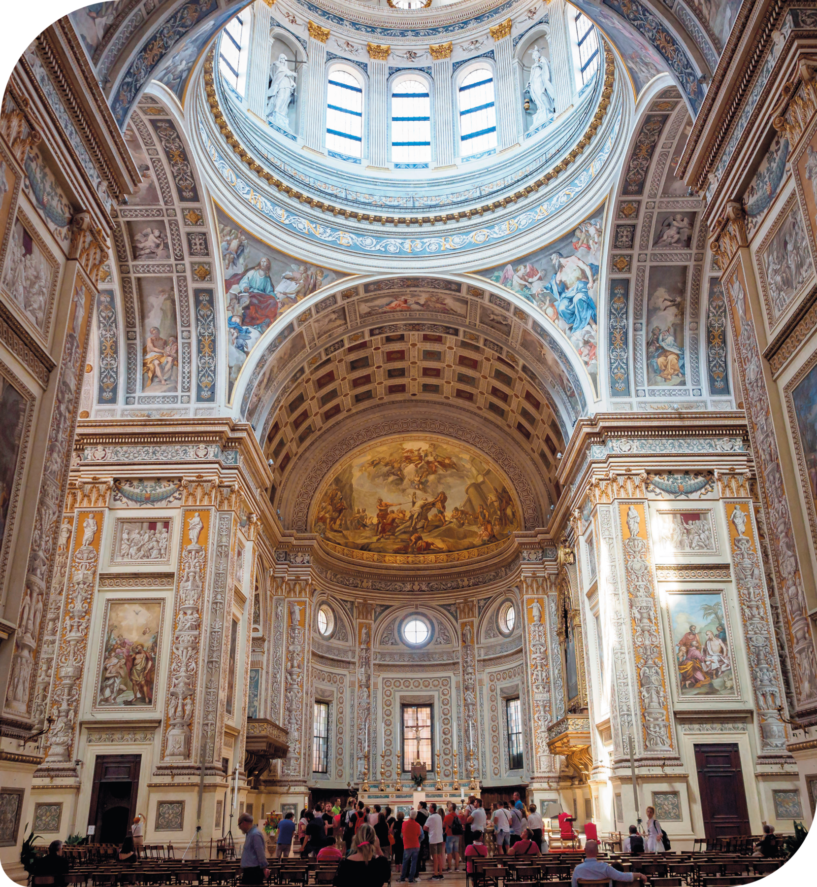 Fotografia. Vista interna de Basílica. Paredes em forma de grandes arcos e decoradas com várias pinturas. Na parte superior, ao centro, uma grande cúpula. Há várias pessoas observando as pinturas nas paredes.
