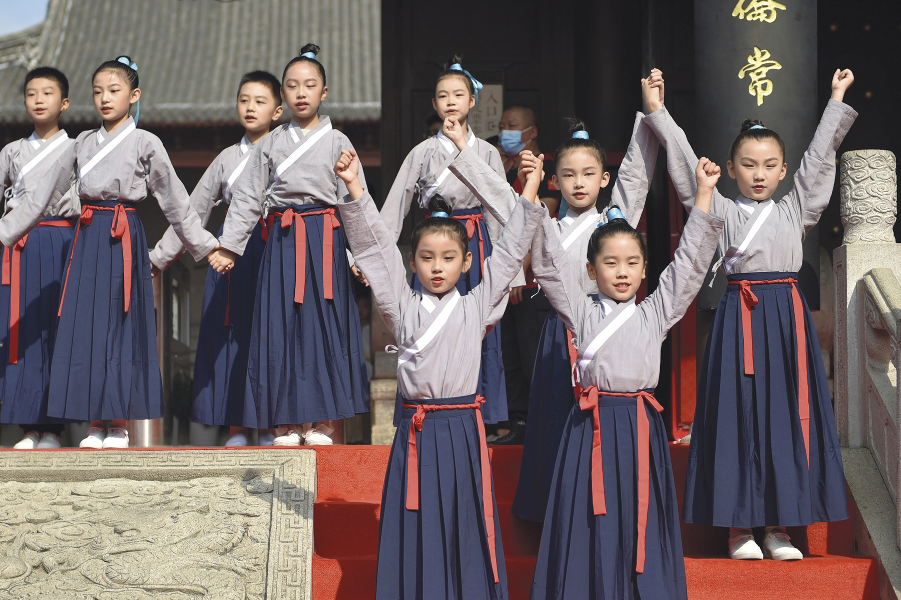 Fotografia. Crianças usando um quimono cinza, saia azul e um tecido vermelho amarrado na cintura. Na parte superior, as crianças estão de mãos dadas com os braços voltados para baixo. Na parte inferior, as crianças estão com os braços estendidos para cima.