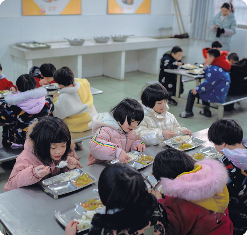 Fotografia. Crianças se alimentando em um refeitório. Elas estão usando casaco, sentadas em um banco. Sobre a mesa, bandejas com arroz e outros legumes.