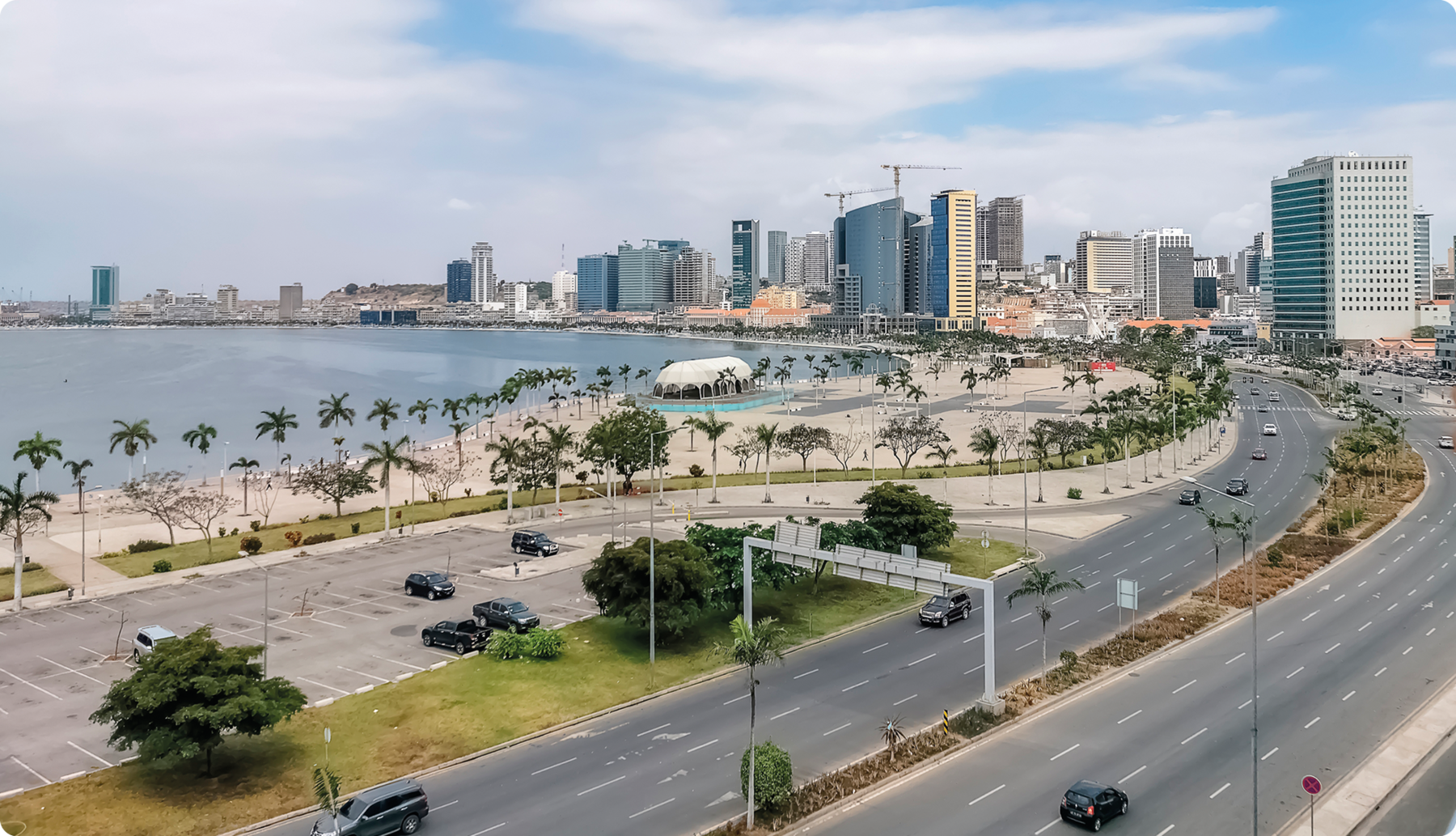 Fotografia. vista de cidade com duas avenidas onde passam alguns carros. À esquerda, orla da praia, várias palmeiras e, ao fundo, o mar. À direita, prédios grandes.