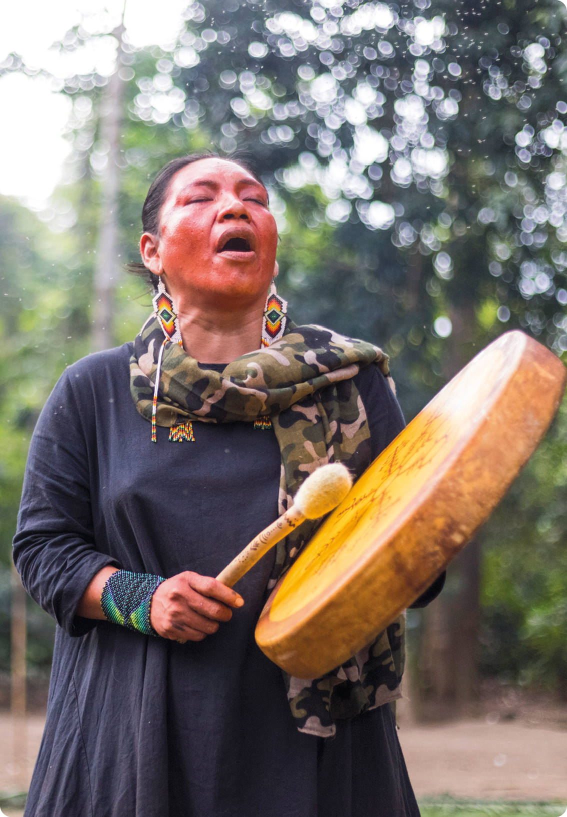 Fotografia. Uma mulher indígena, usando brinco, vestido azul e lenço no pescoço, está de pé, com os olhos fechados, segurando com as mãos um instrumento musical de percussão.