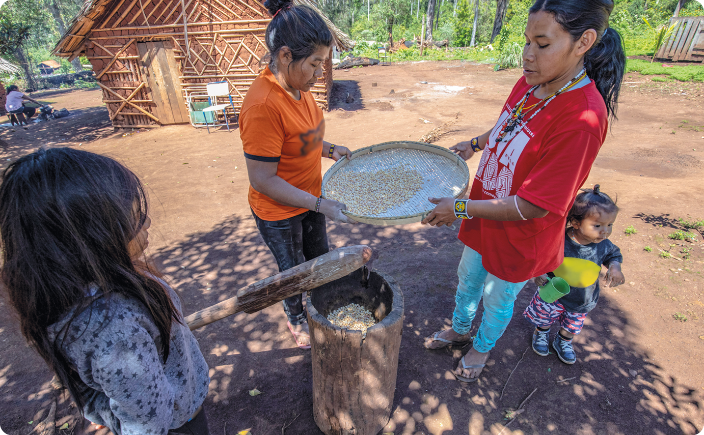 Fotografia. Ao centro, duas mulheres indígenas, usando camisa, calça e adereços no pescoço, estão segurando uma peneira com as mãos na direção de um objeto em formato cilíndrico que está no chão. Perto das mulheres, duas crianças. Atrás, moradia de madeira e vegetação.