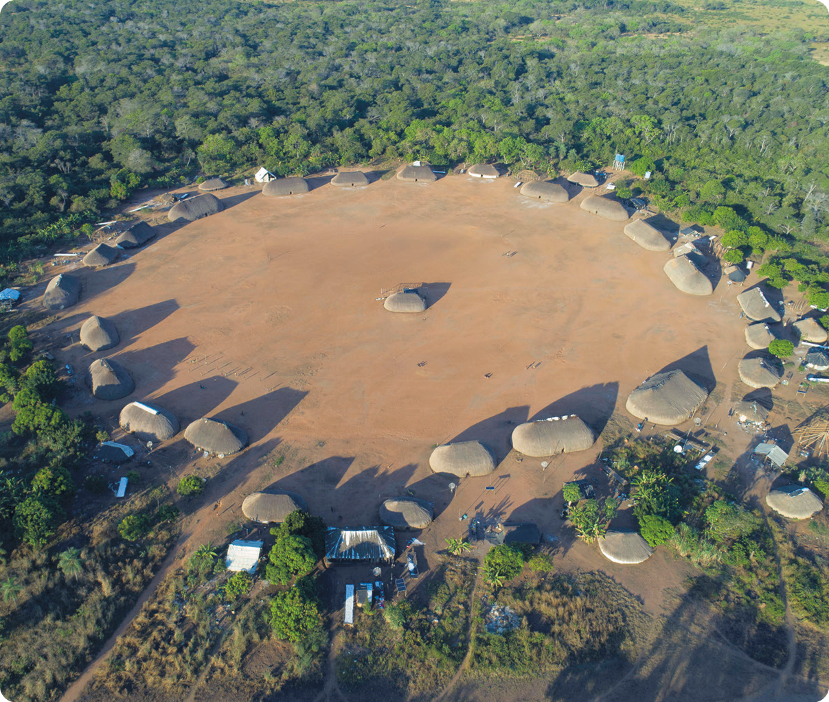 Fotografia. Vista aérea de moradias indígenas dispostas em círculo, com uma moradia no centro. Ao redor, vegetação.
