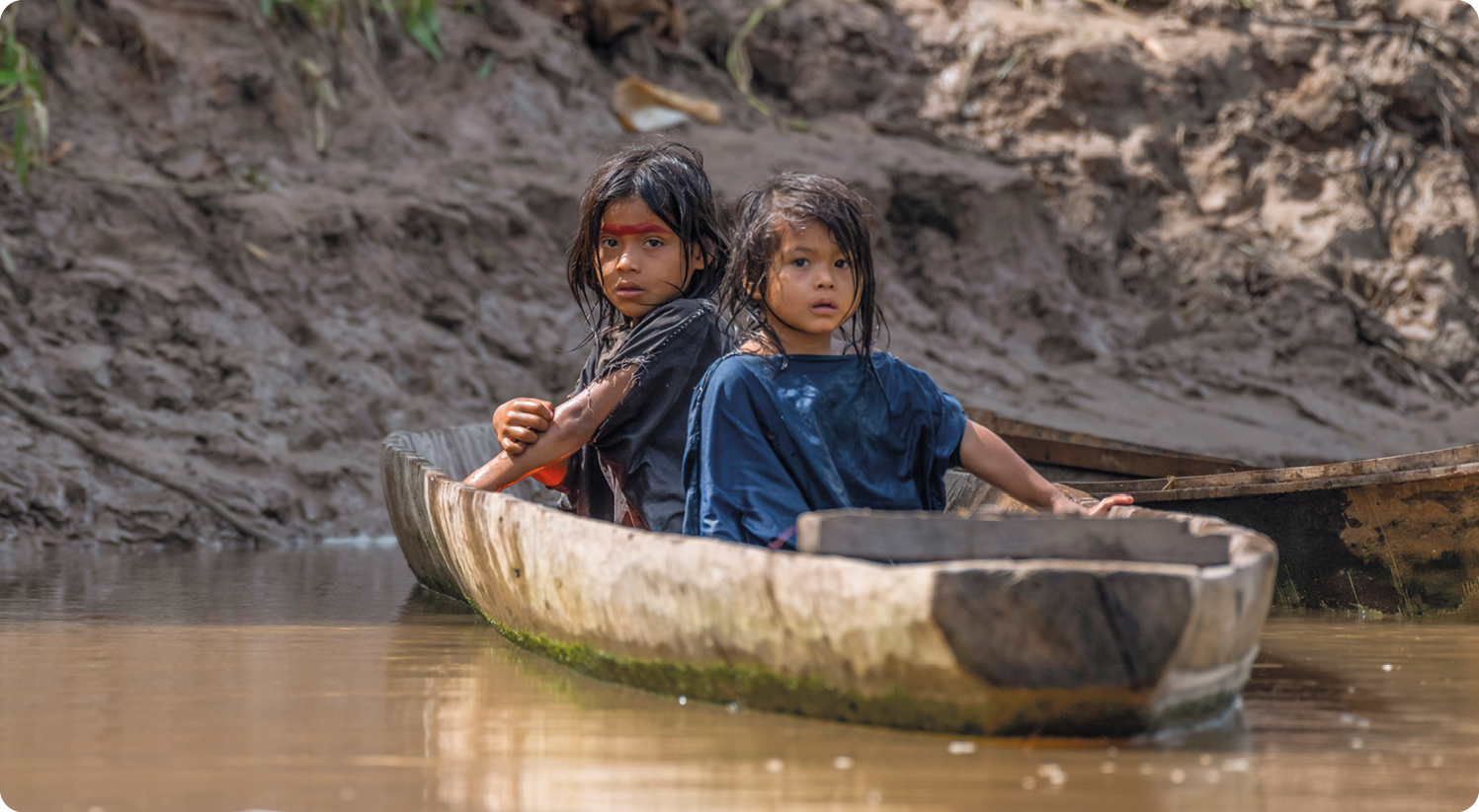 Fotografia. Duas crianças indígenas, usando camiseta, estão dentro de uma pequena embarcação de madeira sobre um corpo de água.