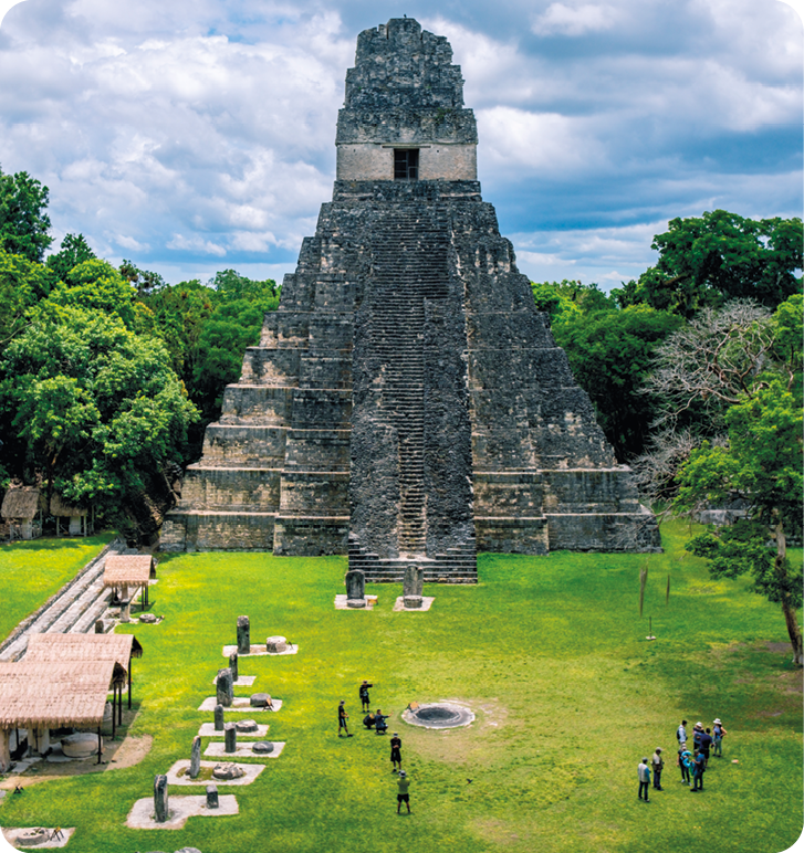 Fotografia. Uma pirâmide de pedra com escada central. Na frente, pessoas caminhando e, em volta, vegetação.