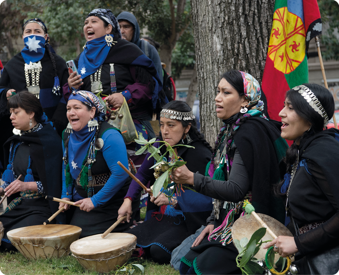 Fotografia. Mulheres indígenas com adorno nos cabelos, usando roupas escuras. Elas estão segurando baquetas sobre tambores, com a boca aberta.