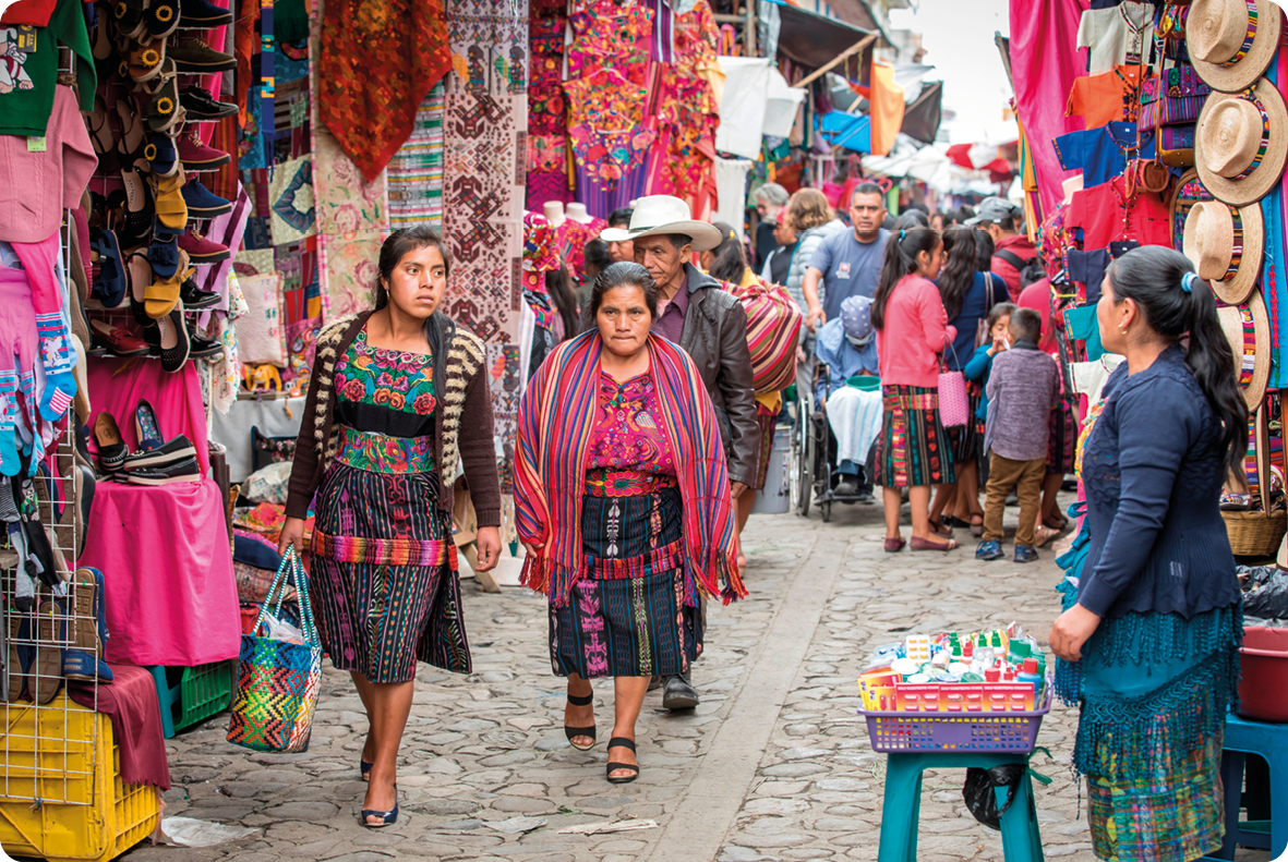Fotografia. À esquerda, duas mulheres, usando roupas coloridas, estão caminhando em uma rua com muitas barracas com mercadorias penduradas, como chapéus e roupas. Ao fundo, pessoas caminhando entre as barracas.