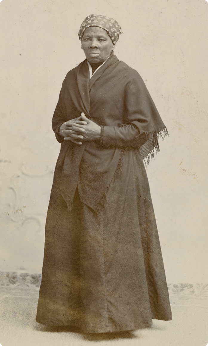 Fotografia em preto e branco. Mulher negra, usando um lenço nos cabelos, vestido, casaco e xale escuro. Ela está de pé com as mãos juntas próximas à barriga.