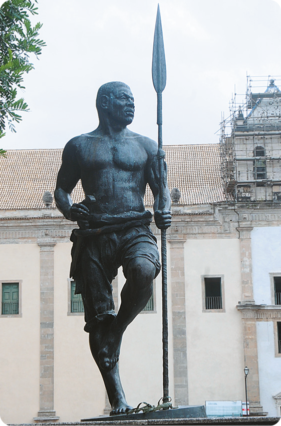 Escultura. Zumbi dos Palmares, homem negro usando short, está descalço com uma perna flexionada e segurando com a mão esquerda uma lança.