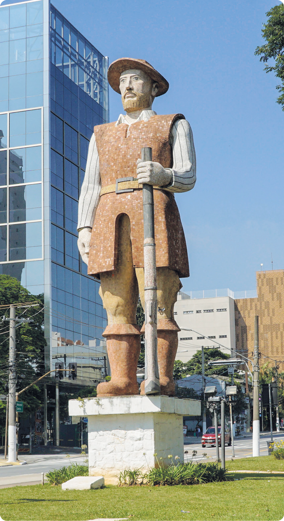 Escultura. Borba Gato, um homem de barba, usando chapéu, camisa, calça e botas, está de pé, segurando com uma das mãos uma arma na posição vertical.