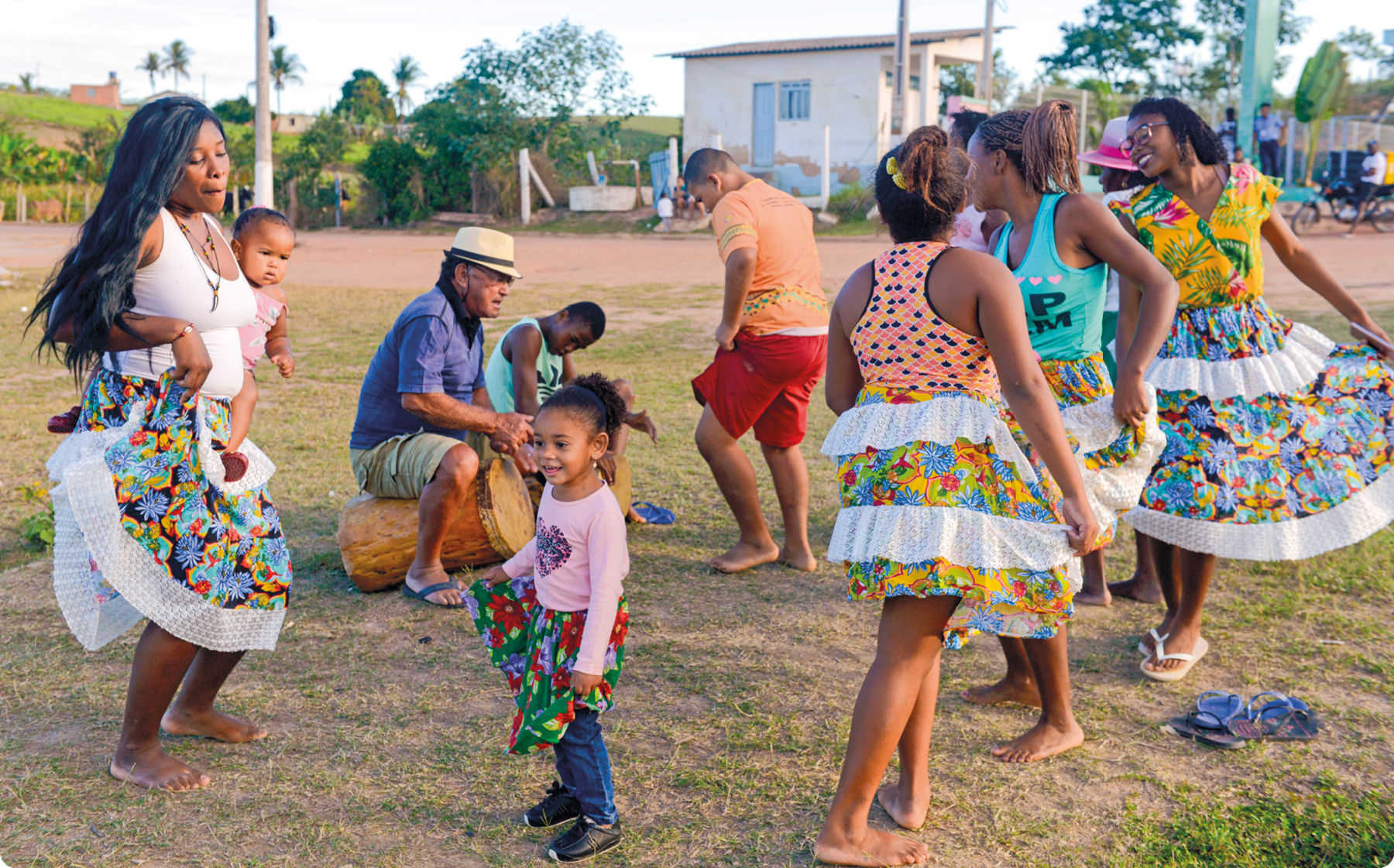 Fotografia. Mulheres e crianças negras, com saias estampadas e coloridas, dançando. Ao fundo, um homem sentado sobre um tambor.