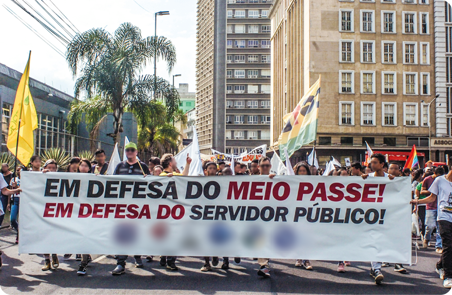 Fotografia. Um grupo de pessoas em uma rua, segurando bandeiras e um cartaz com o texto: EM DEFESA DO MEIO PASSE! EM DEFESA DO SERVIDOR PÚBLICO!
