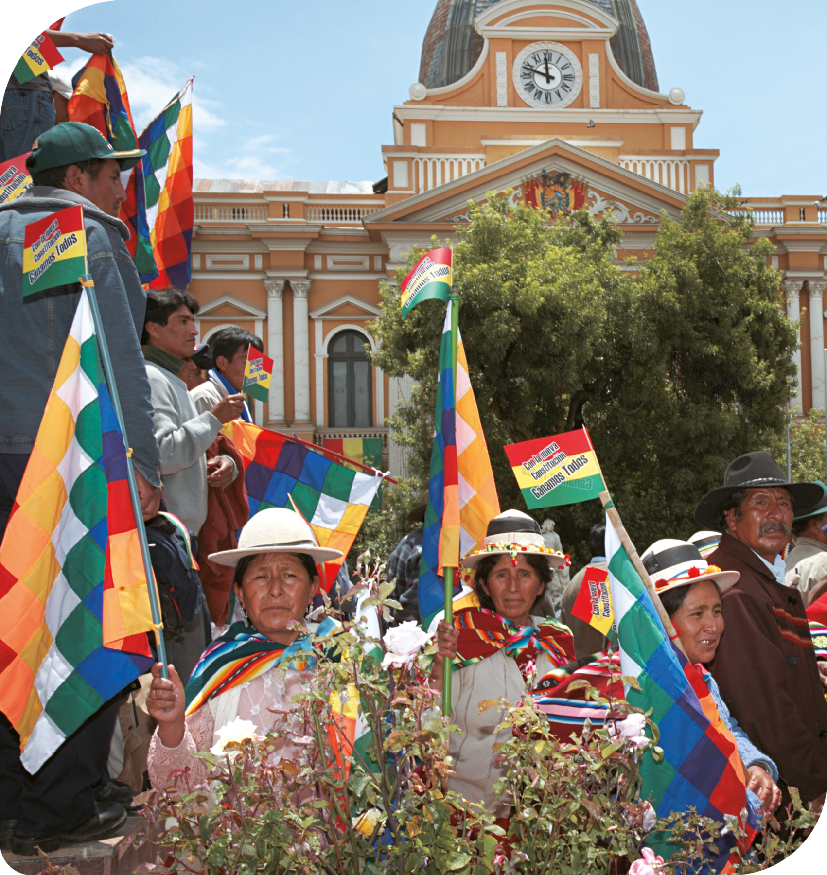 Fotografia. Ao centro, três mulheres usando chapéu, camisa e poncho colorido, estão segurando bandeiras com quadrados coloridos com as mãos. Ao redor, pessoas segurando bandeiras coloridas. Elas estão de pé em uma rua. Atrás, uma construção com muitas janelas.