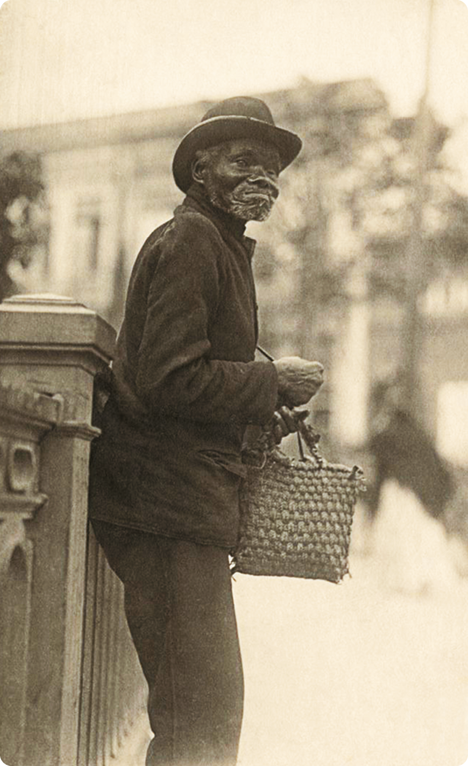 Fotografia em tom sépia. Um homem negro, usando chapéu, camisa e calça, está de pé segurando uma sacola com as mãos.